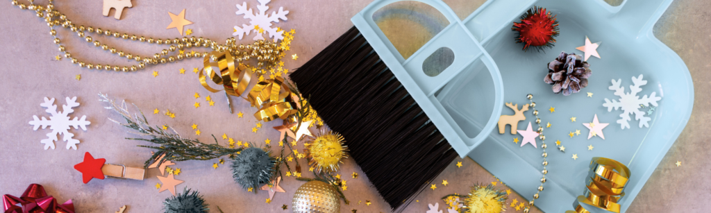 Cleaning tools with festive decorations, including a dustpan and brush, surrounded by holiday-themed ornaments and confetti, symbolizing post-holiday cleaning tips.