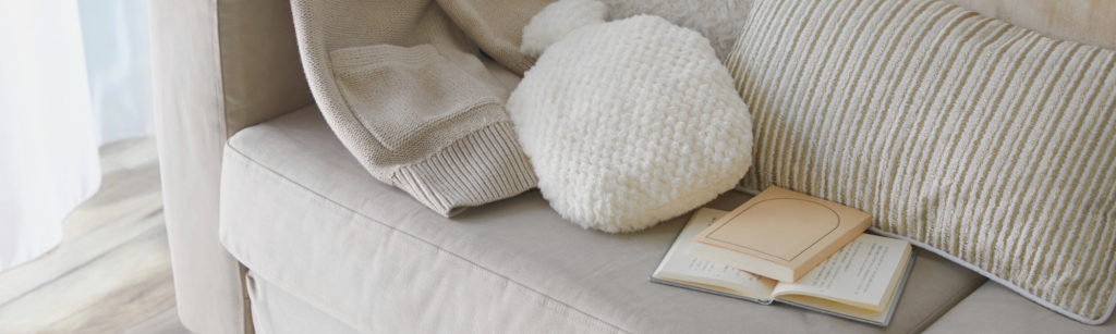 Cozy living room scene featuring a beige sofa adorned with soft blankets and textured pillows, alongside an open book, reflecting themes of home comfort and self-care in cleaning practices.