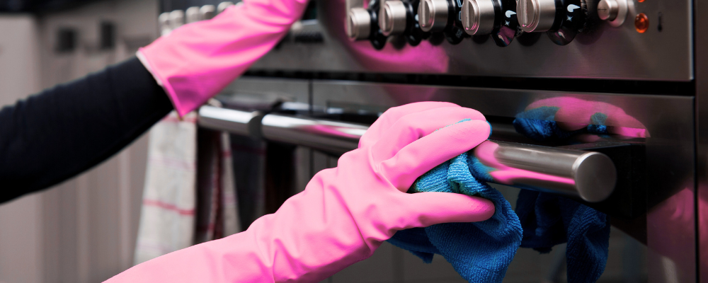 Hands wearing pink rubber gloves cleaning a stainless steel oven handle with a blue cloth, emphasizing preventive commercial cleaning and hygiene practices.
